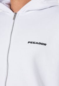 Hvid zip-up hoodie lavet af blødt stof, med en frontlynlås og et sort "PEGADOR" logo på venstre side.