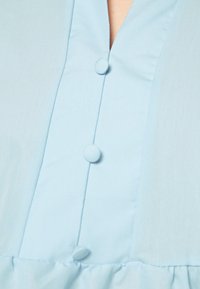 Camisa azul clara com decote em V, três botões combinando na parte da frente e uma bainha de babados franzidos na parte inferior. Textura de tecido suave.