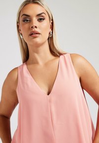 Ljusrosa ärmlös topp med djup V-ringning, med en mjuk, flödande textur och minimalistisk design. Inga synliga mönster eller detaljer.