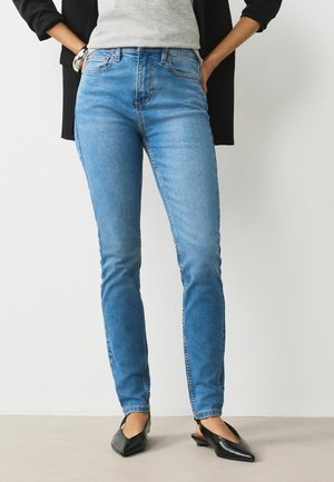 Person iført lyseblå skinny jeans, grå top, sort blazer og sorte spidse slingback sko står med hænderne i lommerne.