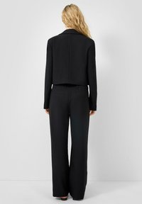 Blazer noir court avec manches longues, coupe cintrée et texture lisse, associé à un pantalon noir à jambes larges avec poches arrière.