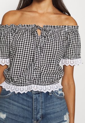 Mujer con blusa de cuadros vichy blanco y negro sin hombros, con borde de encaje y detalle de lazo, combinada con pantalones cortos de mezclilla azul desgastada.