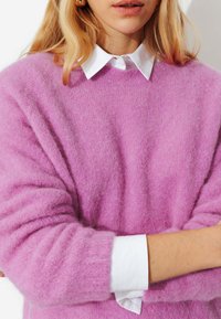 Femme portant un pull lavande doux et duveteux par-dessus une chemise blanche à col, les bras croisés, montrant des boucles d'oreilles dorées et des cheveux blonds.