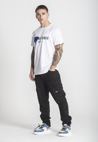 Biele tričko s grafickým potlačou, čierne cargo nohavice a modré tenisky so žltými akcentmi. Pohodlný strih s viditeľnými tetovaniami na rukách.