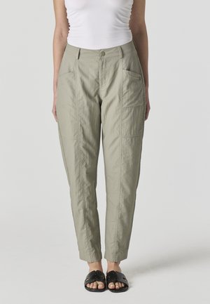 Mujer con pantalones rectos color caqui claro con bolsillos grandes en los costados, una camiseta sin mangas blanca y sandalias negras abiertas de tipo slide.