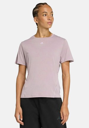 Donna con i capelli intrecciati che indossa una maglietta Adidas color lavanda e pantaloncini neri, guarda verso sinistra su uno sfondo bianco.