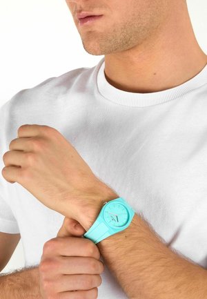 Uomo con camicia bianca che aggiusta un orologio da polso azzurro chiaro al polso sinistro su sfondo bianco.