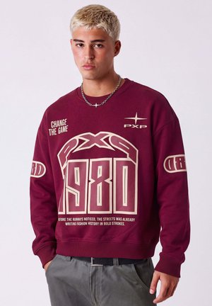 Giovane uomo con capelli corti decolorati che indossa una felpa bordeaux con grande stampa "1980" e pantaloni grigi, in piedi davanti a uno sfondo semplice.