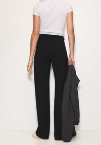 Personne debout de dos, face à un mur, portant un pantalon noir taille haute, un haut blanc à manches courtes, des chaussures blanches, tenant une veste sombre dans la main droite.