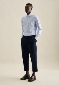 Camicia a righe azzurre e bianche con colletto, abbinata a pantaloni navy plissettati e scarpe nere, su uno sfondo neutro.