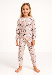 Niña joven de pie descalza, vistiendo un conjunto a juego de sudadera y pantalones rosas cubiertos con corazones blancos contorneados en negro.
