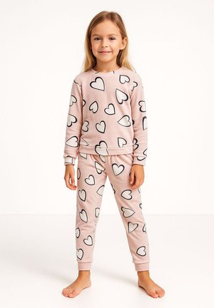 Ein junges Mädchen steht barfuß und trägt einen rosa passenden Sweatshirt und eine Hose, die mit schwarz umrandeten weißen Herzen bedeckt sind.