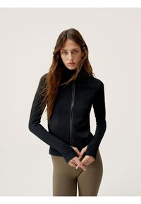 Black athletic zip-up jacket with long sleeves, textured panels, and a high collar, paired with brown fitted leggings.