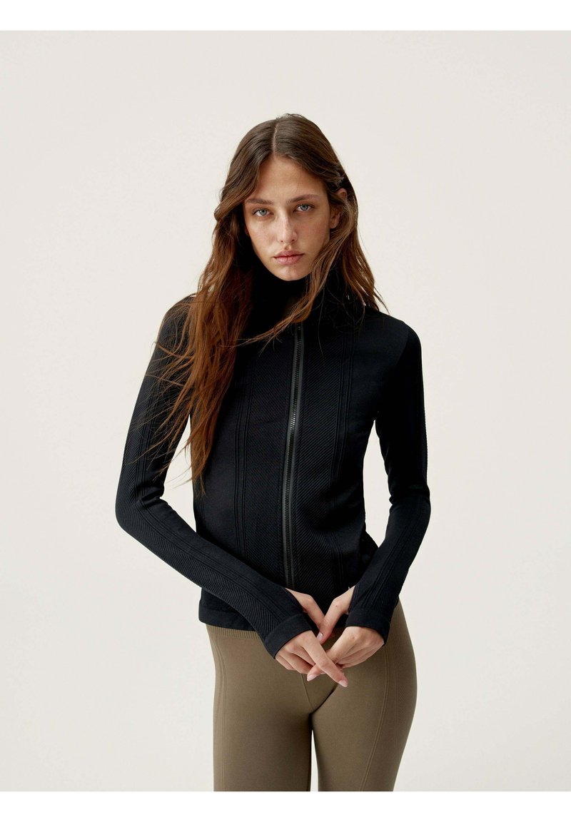 Veste de sport noire à zip, à manches longues, avec des panneaux texturés et un col haut, associée à des leggings marron ajustés.