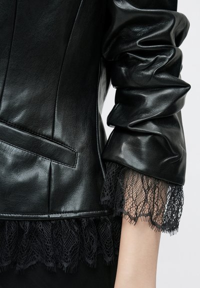 Fekete bőrdzseki, testhez simuló fazonnal, sima textúrával és csipkével díszített ujjakkal. Tartalmaz zseb részletet is.