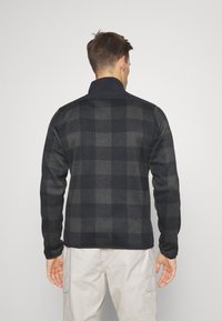 Homme de dos portant une veste à carreaux noire et grise et un pantalon cargo beige clair sur fond uni.