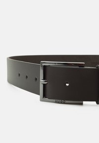 Ceinture en cuir noir avec une boucle rectangulaire en argent, comportant cinq trous pour ajustement et un logo en relief sur la boucle.