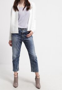 Kvinna klädd i en vit fluffig jacka, lavendel överdel, korta blå jeans och bruna ankellikor stående mot en enkel vit bakgrund.