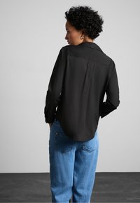 Camicia nera a maniche lunghe con taglio dritto, polsini abbottonati e piega sul retro, indossata con jeans a vita alta blu chiaro.