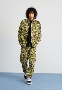 Camo jacket and pants in green and brown fabric. Textured pattern with a relaxed fit. Black shirt underneath and black sneakers.