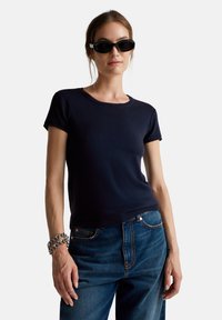 Tyrkysově modré žebrované tričko s krátkým rukávem a kulatým výstřihem, nošené s modrými džínami, doplněné stříbrným řetízkovým náramkem a černými slunečními brýlemi.