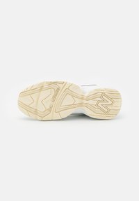 Semelle d'une basket beige avec motifs antidérapants texturés et marquage New Balance visible au talon et au centre.