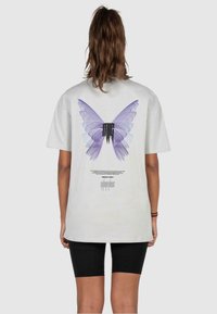 Graues Baumwoll-T-Shirt mit einem großen lila Schmetterlings-Design auf dem Rücken und darunter in schwarzer Schrift. Kurze Ärmel und lockere Passform.