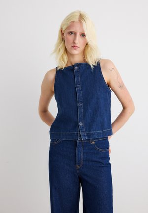 Blondynka o włosach sięgających do ramion, ubrana w bezrękawnikowy, zapinany na guziki ciemnoniebieski top jeansowy i pasujące do niego jeansy z wysokim stanem, stojąca na jednolitym tle.