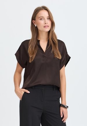 Femme aux longs cheveux châtain clair, portant un chemisier ample marron foncé et un pantalon noir, debout avec une main dans la poche et une expression neutre.