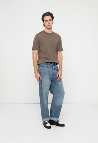 Brun t-skjorte med korte ermer, løse blå jeans i denim med oppbrettede mansjetter, og svart-hvite sko, mot en ensfarget bakgrunn.