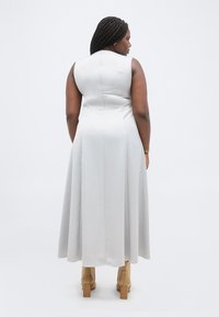 Γυναίκα με πλεξούδες, φορώντας ένα αμάνικο γκρι μίντι φόρεμα και ταμπά μποτάκια με τακούνι, στέκεται με την πλάτη προς το φακό σε λευκό φόντο.