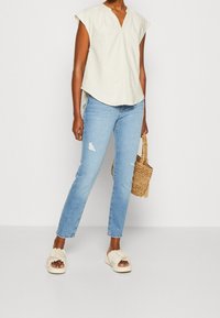Top beige claro de manga corta con escote en V, combinado con jeans destrozados azul claro. El modelo lleva una bolsa de mimbre y calza zapatos tipo slip-on en color crema.