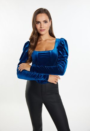 Blauwe fluwelen top met lange mouwen, een vierkante halslijn en geplooide pofmouwen, gecombineerd met zwarte strakke broek. Gladde textuur met een subtiele glans.