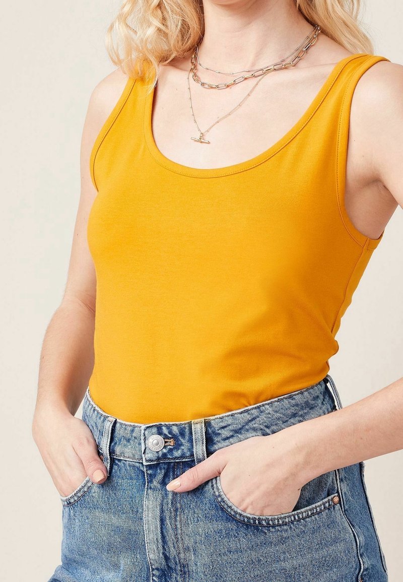 Débardeur jaune en coton, avec un col rond plongeant et des bretelles épaisses. Associé à un short en denim bleu taille haute.