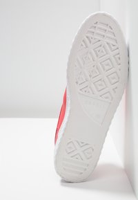 Semelle d'une basket rouge Esprit avec un motif de bande de roulement blanc détaillé reposant sur une surface blanche contre un mur blanc.