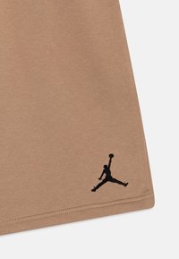 Hnědá bavlněná látka s hladkou texturou, s černým vyšitým logem Jumpman blízko pravého dolního rohu.
