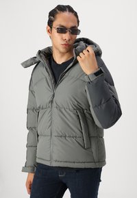 Grå pufferjakke med zip foran, hætte og tekstureret finish. Features inkluderer en blanding af mørke og lyse grå sektioner samt sidelommer.