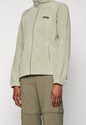 Osoba ubrana w jasnozieloną kurtkę polarową Columbia z zamkniętą kieszenią na zamek oraz oliwkowozielone spodnie, stojąca na jednolitym tle.