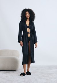 Μαύρη σατέν ρόμπα με μακριά μανίκια, ζώνη και V-λαιμόκοψη. Φοριέται πάνω από αντίστοιχο μαύρο εσώρουχο. Μαλακή υφή, κομψός σχεδιασμός.