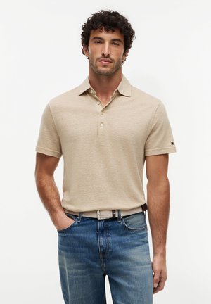 Mand iført beige kortærmet polo og blå jeans med beige flettet bælte, stående foran en ensfarvet hvid baggrund.