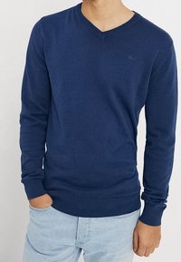 Man wearing a navy blue V-neck sweater with ribbed cuffs and hem, paired with light blue jeans.