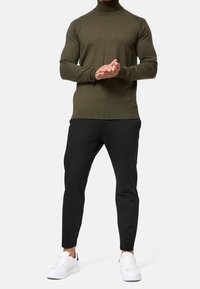 Olijfgroen gebreide coltrui met ribgebreide onderkant en mouwen, gecombineerd met zwarte slim-fit broeken en witte sneakers.