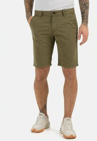 Olivegrüne Baumwollshorts mit Reißverschluss und Knopfverschluss, flachem Frontdesign, Seitentaschen und umgeschlagenen Säumen. Kombiniert mit weißen Sneakers.