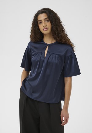 Kobieta z długimi kręconymi włosami, ubrana w granatową satynową bluzkę z krótkim rękawem i czarne szerokie spodnie, stojąca z jedną ręką w kieszeni.