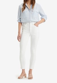 Camisa de botones a rayas azul claro con mangas remangadas, combinada con jeans blancos ajustados y sandalias de tacón nude.