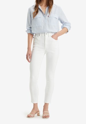 Camisa de botones a rayas azul claro con mangas remangadas, combinada con jeans blancos ajustados y sandalias de tacón nude.