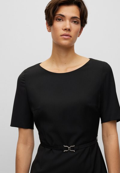 Robe noire ajustée avec des manches courtes, un col rond et une ceinture ornée de détails métalliques à la taille, texture de tissu lisse.