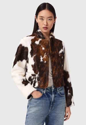 Femeie cu păr drept și închis la culoare, purtând o jachetă blană cu imprimeu de vacă maro și alb și blugi albaștri, cu o mână în buzunar, fundal neutru.