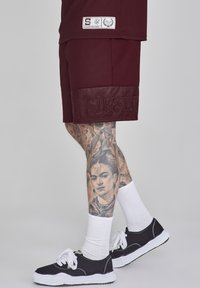 Burgundské športové šortky s sieťovinovou textúrou a reliéfnym logom "SIKSILK" na leme. Nosí sa s čiernymi teniskami a bielymi ponožkami.