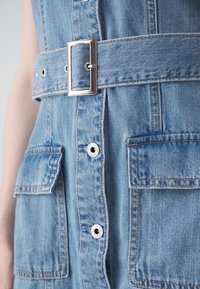 Abito in denim azzurro chiaro con vita cinturata, dotato di fibbia metallica, due tasche frontali e chiusura a bottoni con dettagli in hardware argentato.